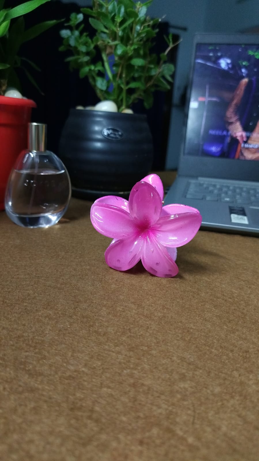 Pink Hawaiian flower hair clip by Serraw placed on a brown surface near a laptop and perfume bottle